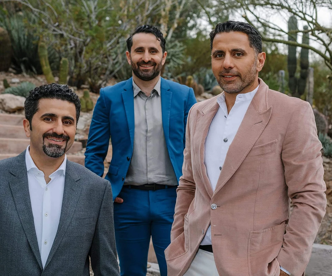 A group portrait of three men, including all Dr. Torabi in a bright blue suit, standing outdoors in a desert landscape with cacti in the background. They are dressed in professional blazers and slacks, smiling at the camera.
