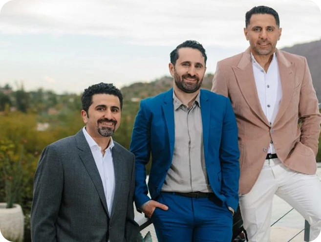 A group portrait of three men, including all Dr. Torabi in a bright blue suit, standing outdoors in a desert landscape with cacti in the background. They are dressed in professional blazers and slacks, smiling at the camera.