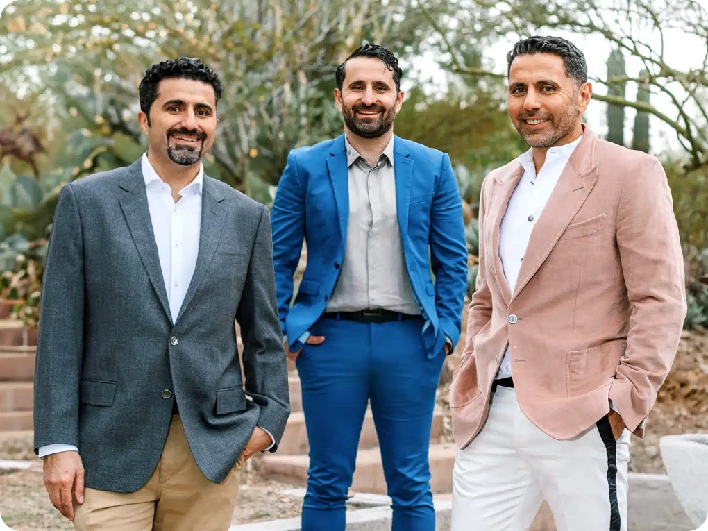 A group portrait of three men, including all Dr. Torabi in a bright blue suit, standing outdoors in a desert landscape with cacti in the background. They are dressed in professional blazers and slacks, smiling at the camera.