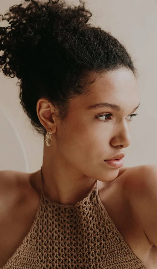 A profile portrait of a woman with curly hair tied back, wearing gold hoop earrings and a tan crochet top. She is looking to the side, highlighting her facial profile and smooth skin.