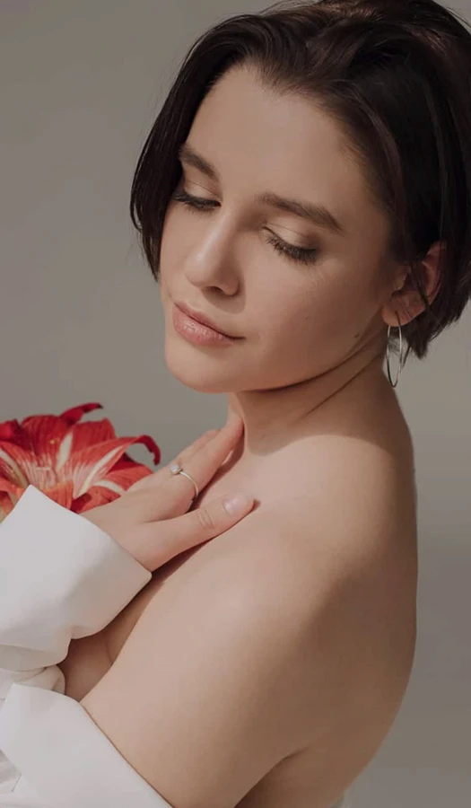 A soft-focus portrait of a woman with short dark hair, looking downward with a peaceful expression. She is draped in a white garment and holds a vibrant red flower near her shoulder.