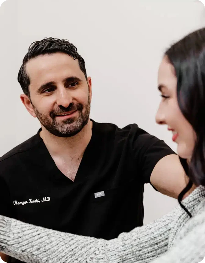 A close-up, warm interaction showing Dr. Ramyar Torabi, MD, wearing black scrubs, smiling kindly at a female patient who is partially visible in the foreground.