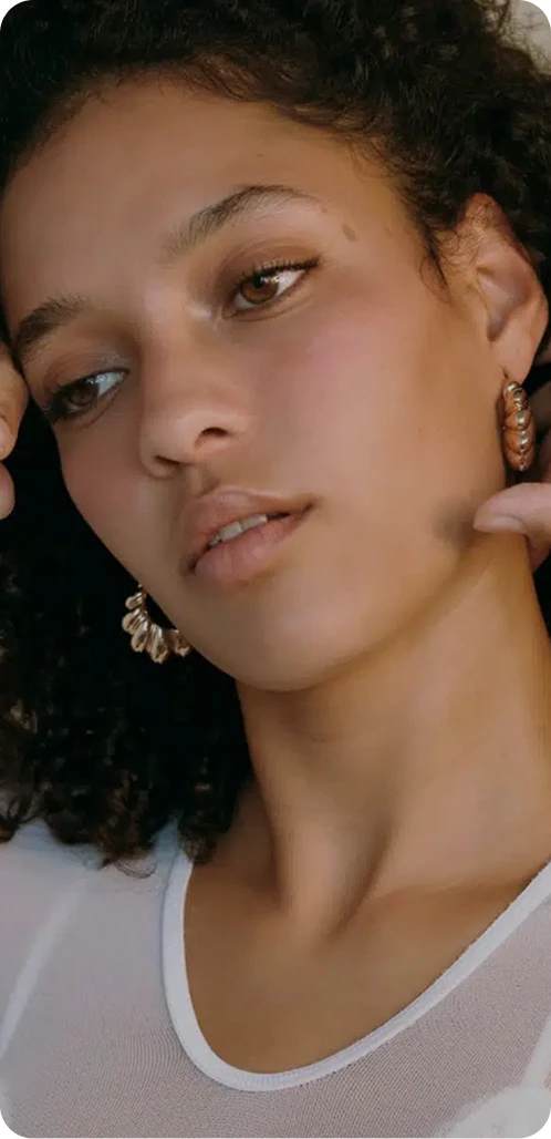 A striking close-up of a woman with curly hair and brown eyes. She is wearing gold hoop earrings and looking slightly off-camera, emphasizing smooth skin and facial symmetry.