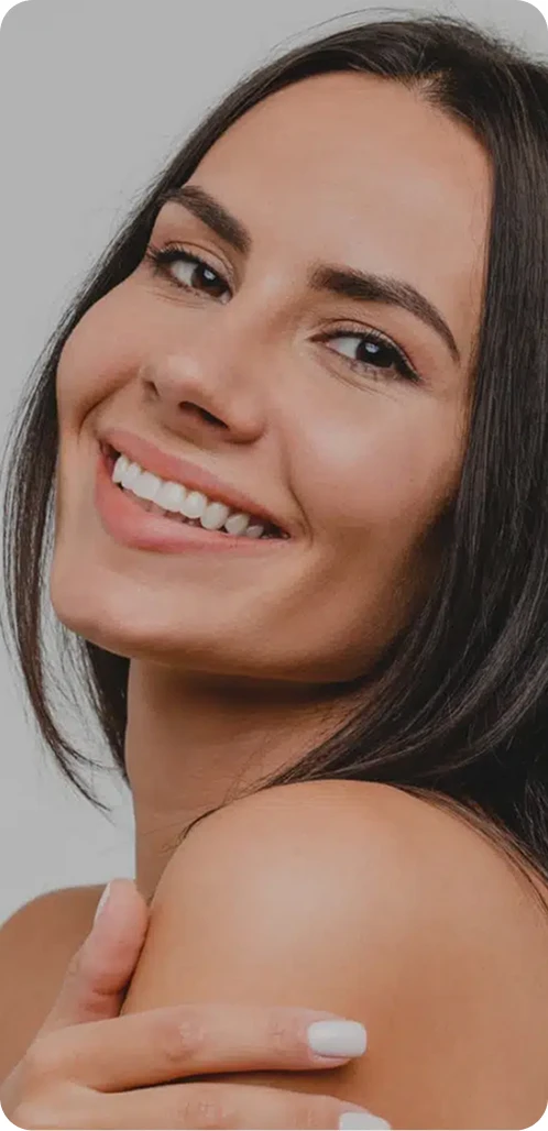 A close-up, high-angle portrait of a woman with dark hair and a bright smile, resting her hand on her shoulder and looking into the camera.