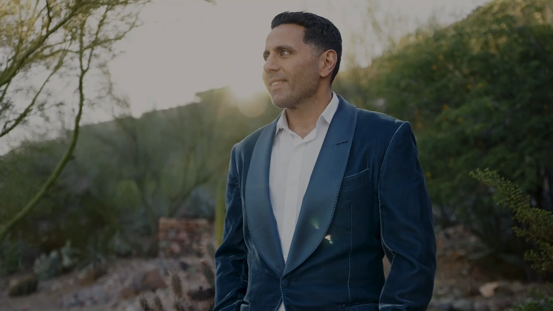 A portrait of Dr. Torabi wearing a textured blue velvet blazer over a white shirt, looking thoughtfully off to the side. The background shows a soft-focus desert sunset with glowing natural light.
