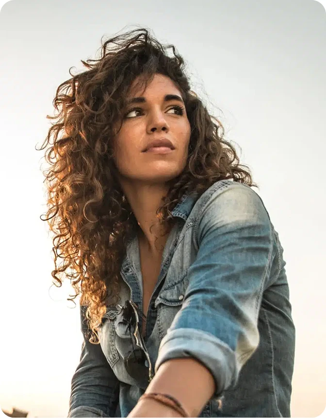 A woman with voluminous, curly brown hair and a denim shirt looks off into the distance against a bright, sunlit outdoor background.