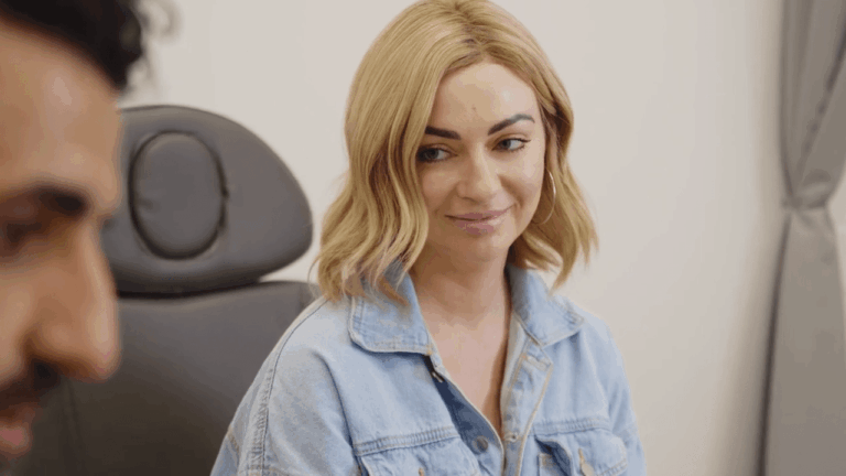 A blonde woman in a denim jacket smiles subtly during a conversation in a medical office. Dr. Torabi's profile is partially visible in the blurred foreground.