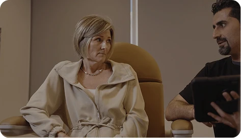 Dr. Torabi, visible in profile, holds a tablet and speaks with a mature female patient during a consultation. The patient, wearing a beige jacket, listens intently.
