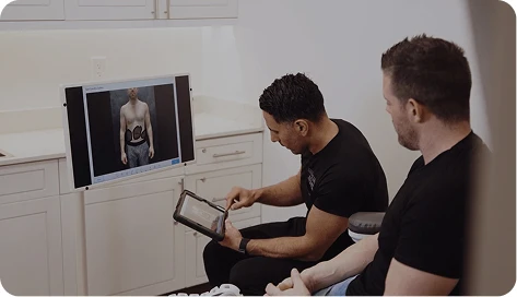 Dr. Torabi sits with a male patient in a consultation room. Dr. Torabi is using a tablet to demonstrate a procedure, which is mirrored on a large monitor showing a 3D medical body scan.