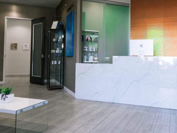 A view of a modern medical office reception area. A large, white marble-patterned reception desk stands in the foreground, with a white computer monitor visible behind it. To the left, a dark wood door with a frosted glass pane is slightly open. A tall glass display case stands next to the door, filled with various white bottles and professional products. A blue abstract painting is mounted on the taupe wall, and the floor is a light grey wood-plank tile. - in Arizona