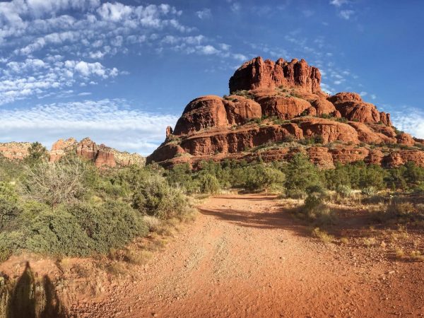 A wide-angle shot of a dusty, reddish-brown dirt path leading toward a massive red rock butte under a bright blue sky. The rock formation is tiered with jagged edges and sparse greenery clinging to its crevices. Thick, green desert shrubs and small trees line both sides of the trail. The sky is decorated with a unique pattern of small, white "popcorn" clouds that stretch toward the horizon. In the bottom left corner, the faint shadows of the photographer are visible on the ground. - Arrowhead in Arizona