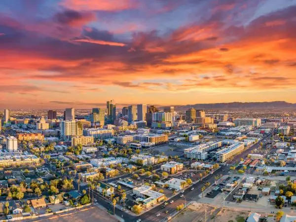 An expansive aerial view of a sprawling metropolitan skyline during a vibrant sunset. The sky is filled with wispy, elongated clouds catching shades of deep orange, pink, and purple. In the center, a dense cluster of modern glass and concrete skyscrapers rises above the surrounding urban landscape. The foreground consists of lower-density commercial and residential buildings, gridded streets with sparse evening traffic, and patches of green trees. In the far distance, purple-toned mountain ranges sit beneath the hazy horizon under a soft golden glow.- Phoenix in Arizona