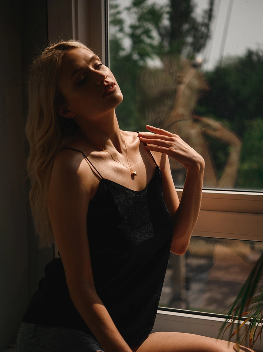 A blonde woman wearing a black silk camisole and a gold heart necklace sits by a window. She is posed with one hand near her collarbone, emphasizing a graceful, proportional upper body and a natural, lightened chest silhouette. - Breast Reduction in Arizona