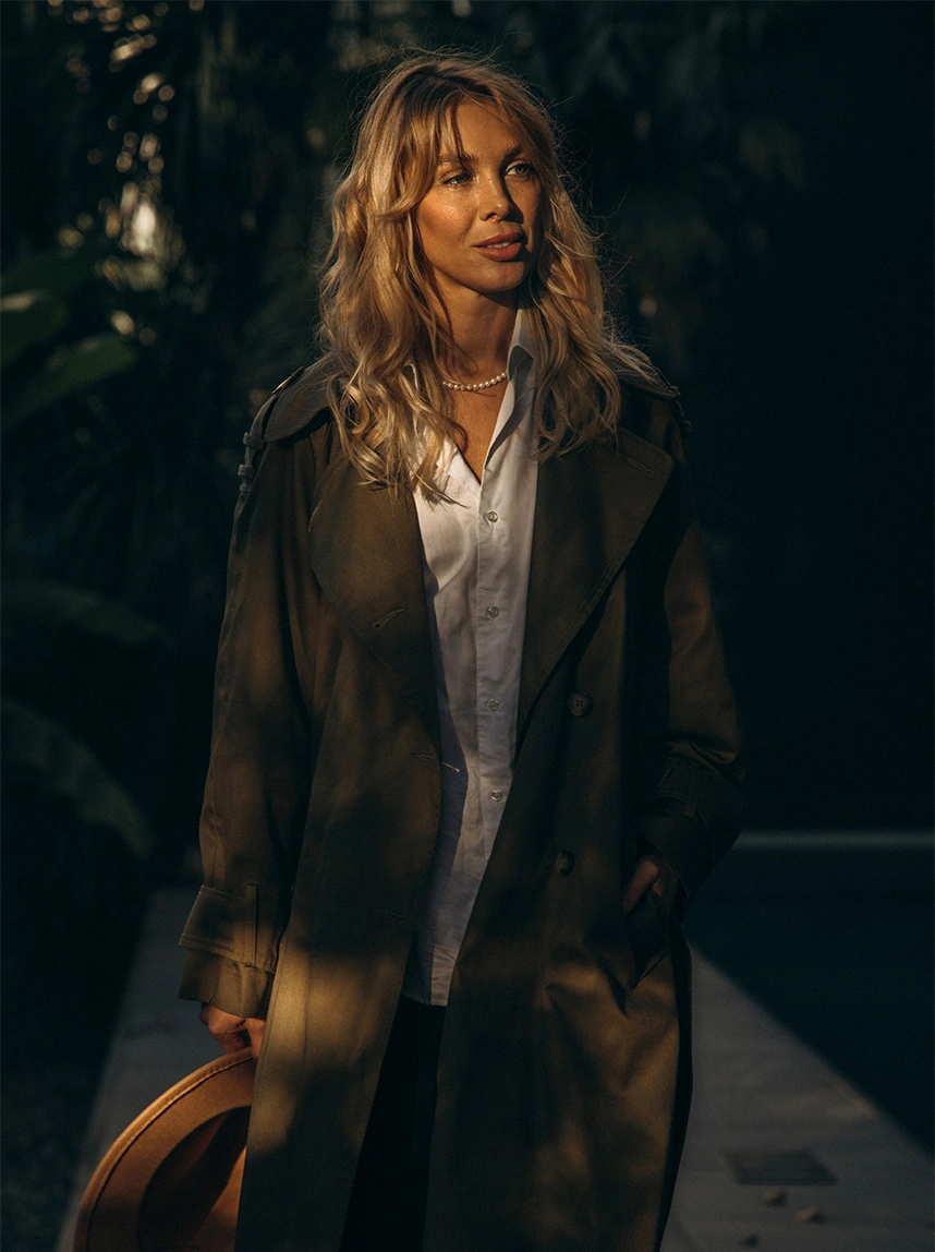 A woman with blonde wavy hair wearing a white button-down shirt and an olive trench coat stands outdoors in soft evening light, representing wellness and lifestyle confidence. - Emsella in Arizona