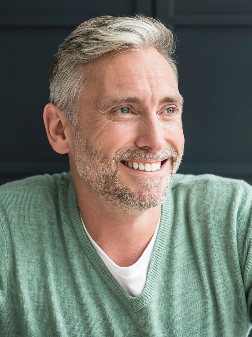 A smiling man with graying hair and a green V-neck sweater looks off-camera, showcasing a refreshed, alert appearance with well-positioned brows. - Eyebrow Ptosis Repair in Arizona