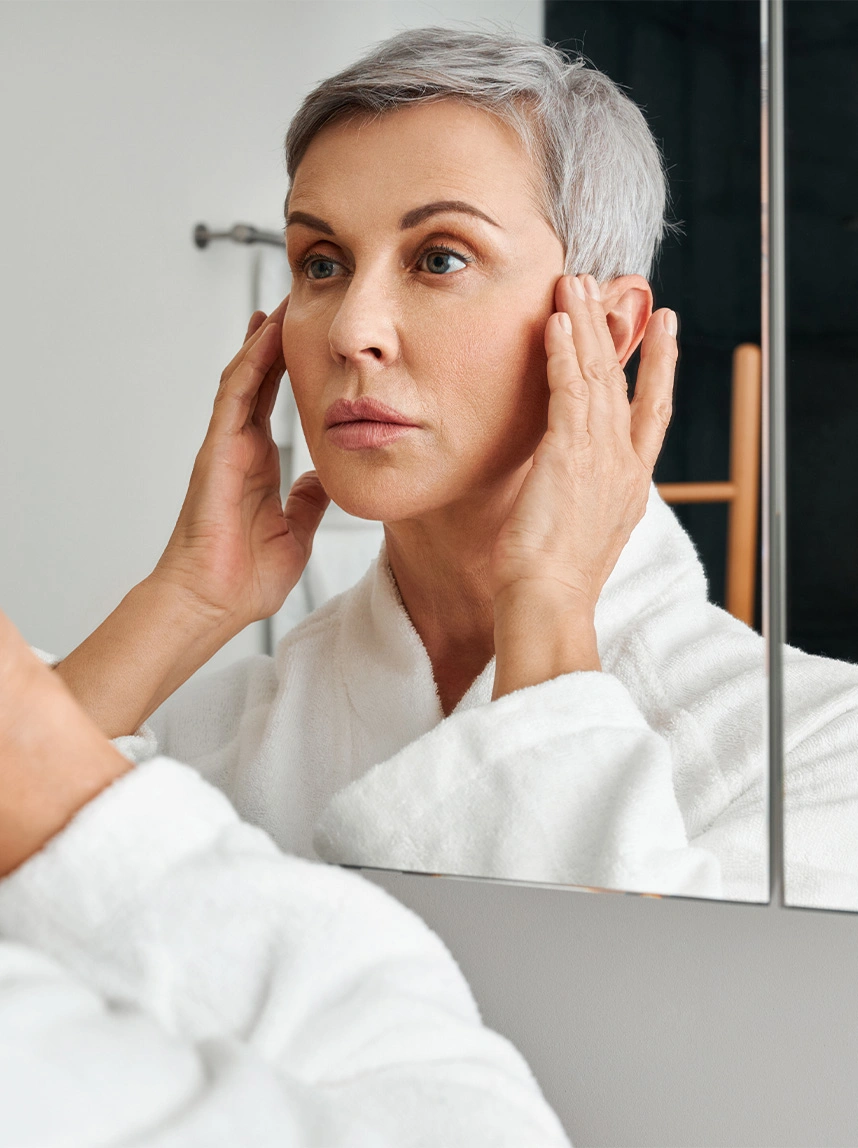 A woman with short silver hair in a white bathrobe looks into a mirror while touching her temples, illustrating a clear and healthy periocular (eye area) skin surface. - Eyelid Lesions in Arizona