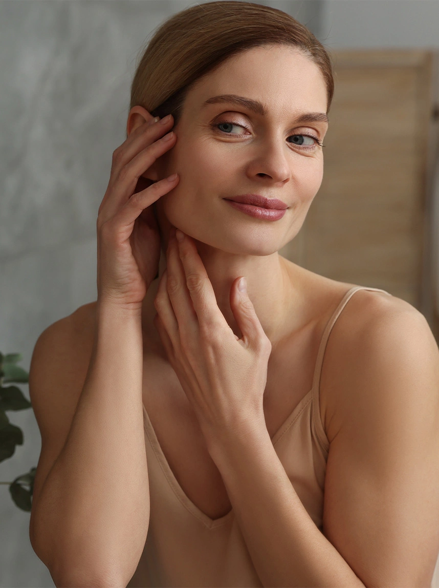 A woman in a tan camisole touches her face gently, showcasing properly aligned eyelids and a symmetrical, comfortable eye expression. - Eyelid Misplacement in Arizona