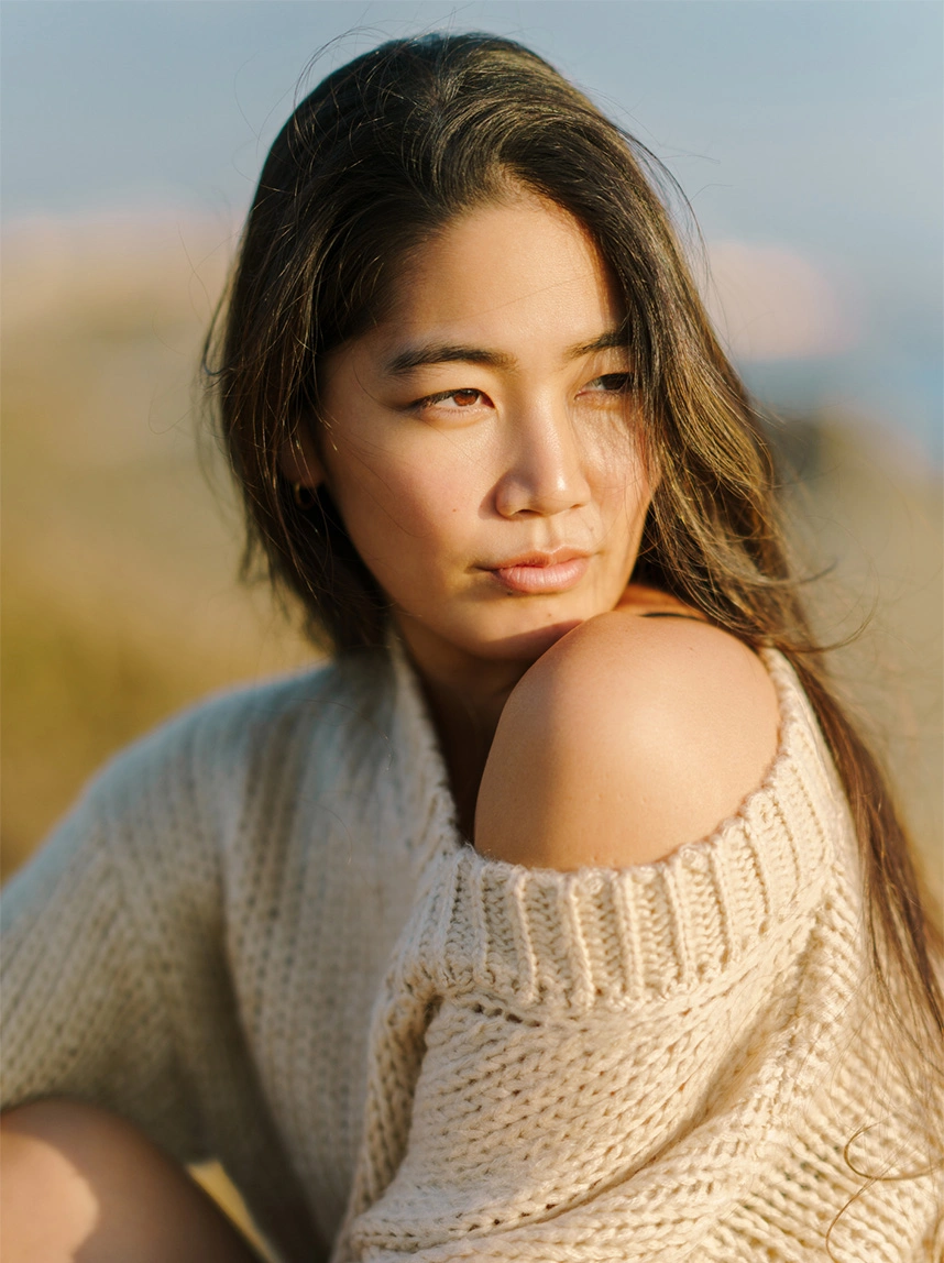 A woman with dark hair wearing a cream-colored chunky knit sweater looks over her shoulder. The soft outdoor lighting highlights the natural, youthful volume and smooth contours of her cheeks and mid-face. - Facial Balancing Fat Transfer in Arizona