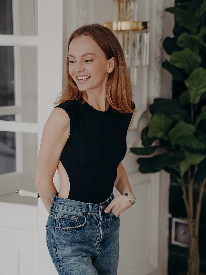 A woman with strawberry blonde hair smiles brightly while standing in a stylish interior, wearing a black backless bodysuit and jeans, radiating vitality and energy. - Hormone Replacement in Arizona