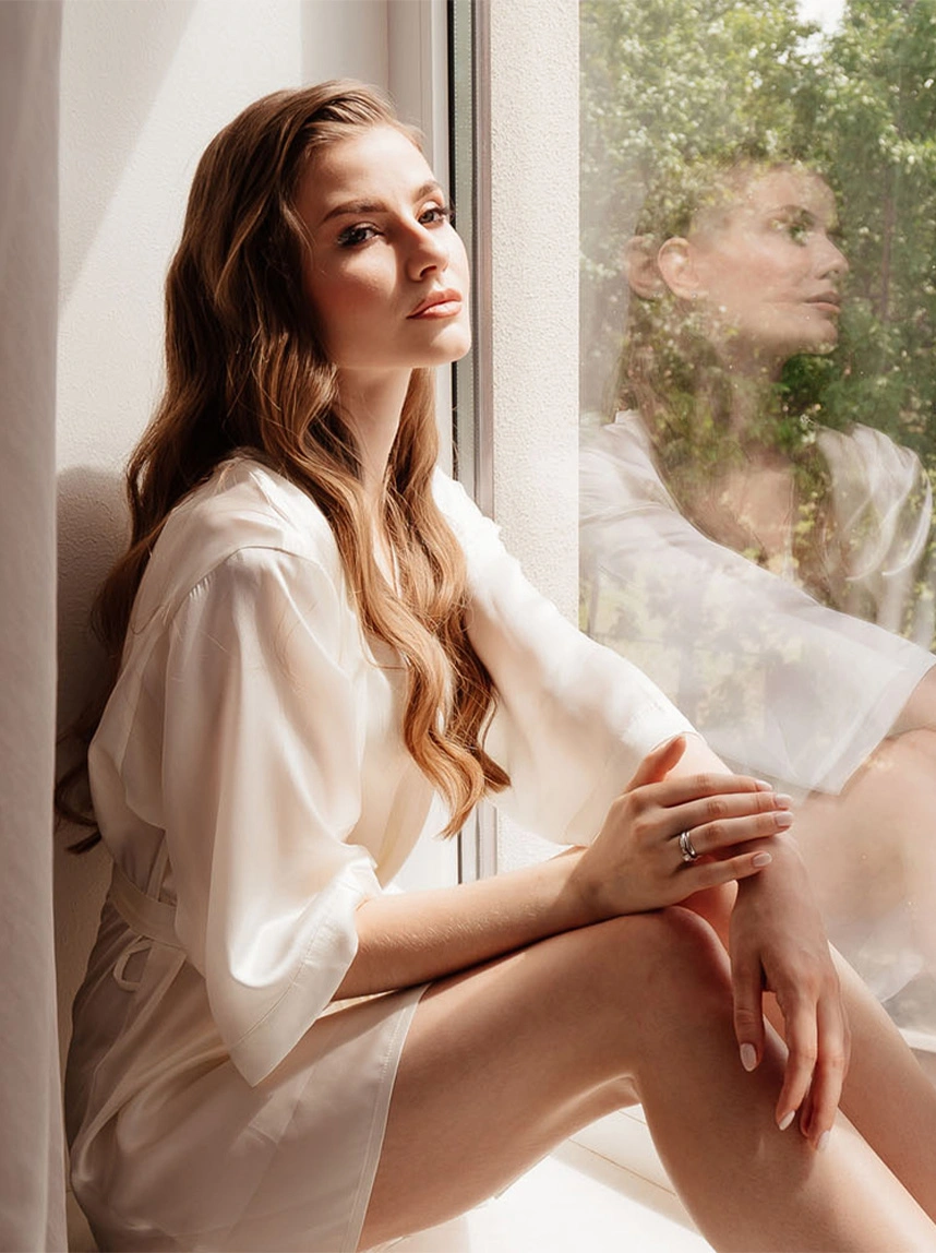 A woman in a white silk robe sits by a window in a soft, bright room, representing a sense of personal comfort and intimate aesthetic confidence. - Labiaplasty in Arizona