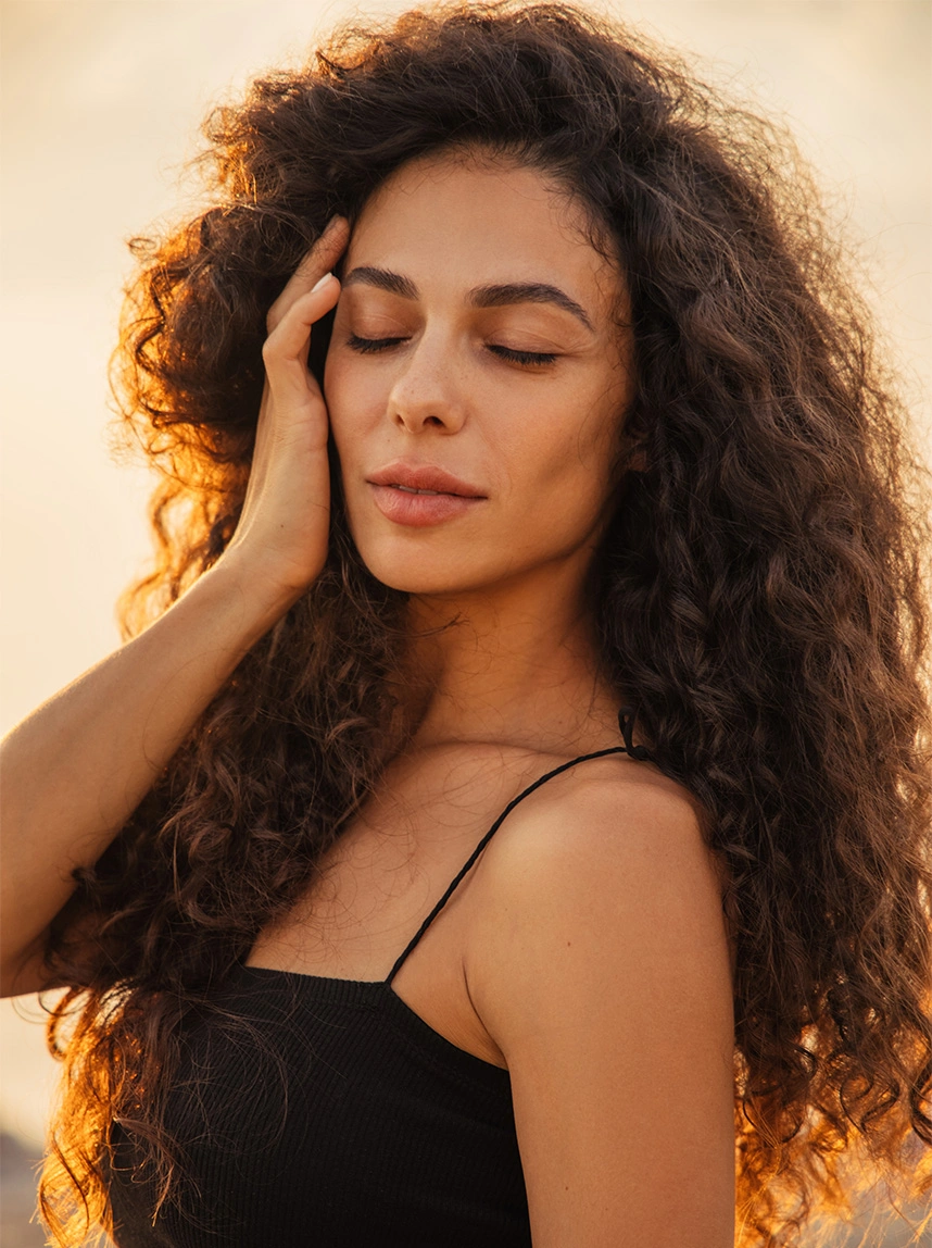 A woman with voluminous dark curls closes her eyes in golden-hour sunlight, showcasing a radiant, even skin tone and healthy complexion. - Lasers in Arizona