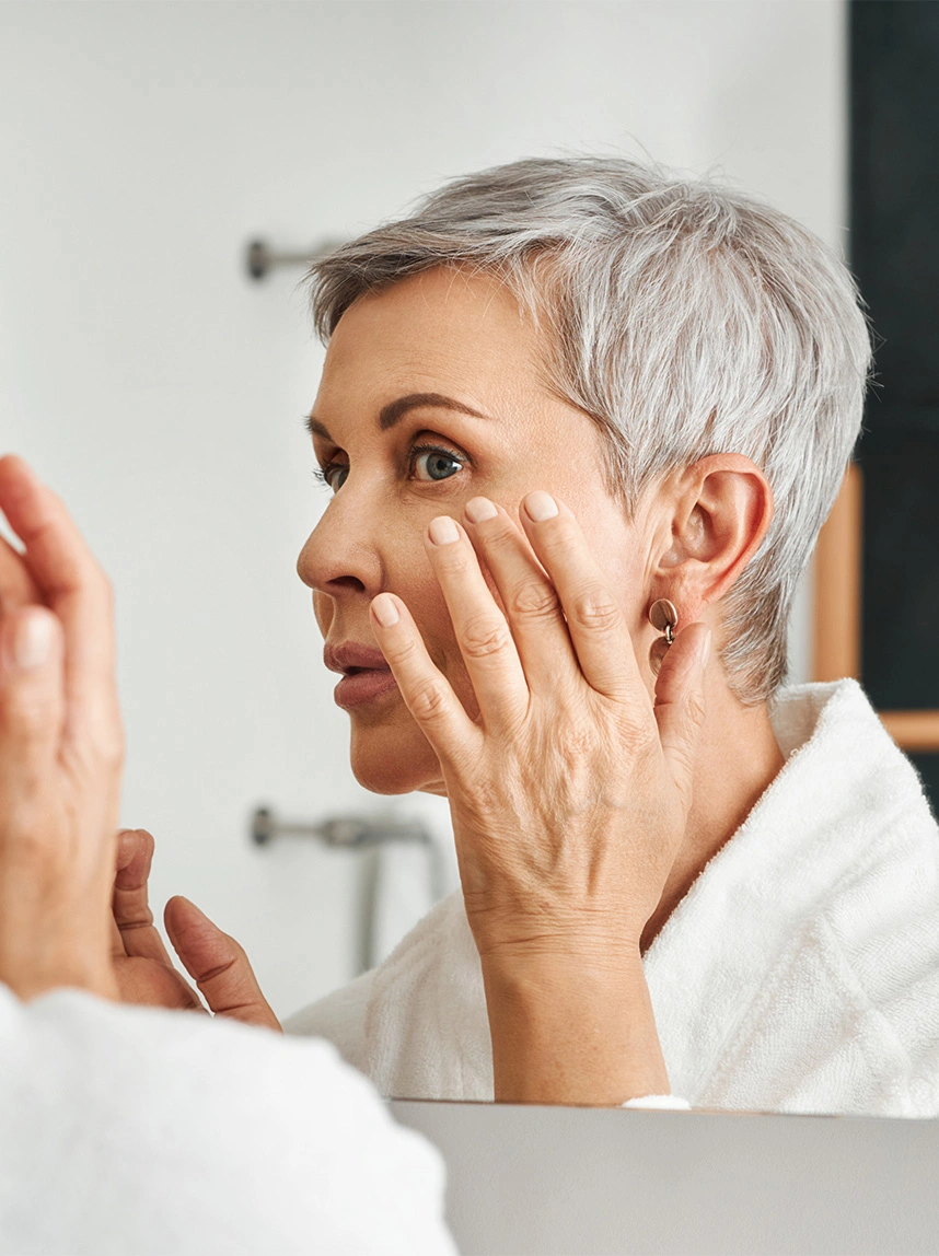 A woman with short silver hair looks into a mirror, gently touching the skin near her eye to inspect a clear, healthy complexion and restored skin texture. - Mohs Reconstructive Surgery in Arizona