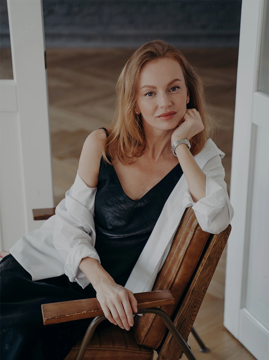 A woman with strawberry-blonde hair wearing a black camisole and a white unbuttoned shirt sits in a leather chair, resting her chin on her hand to highlight a smooth, defined neck and jaw area. - Neck Lift in Arizona