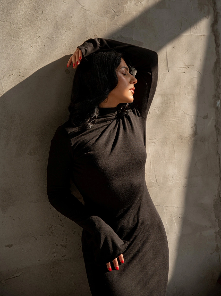A woman in a long-sleeved black turtleneck dress poses against a neutral wall. The directional sunlight emphasizes a sleek, tightened body contour and refined silhouette. - Renuvion in Arizona
