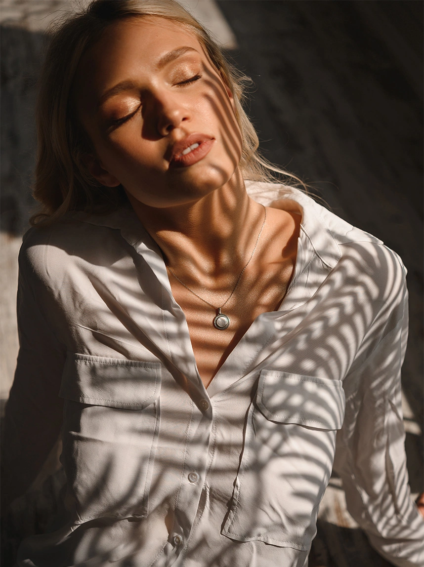 A woman with blonde hair wearing a white button-down shirt and a silver pendant necklace closes her eyes in dappled sunlight, representing a refreshed and rejuvenated skin appearance. - Renuvion in Arizona