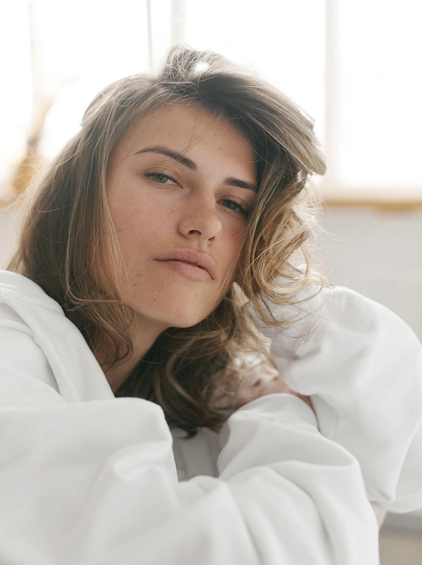 A woman with wavy hair wearing a white hoodie looks into the camera with a relaxed expression, showcasing a natural-looking and well-proportioned nose that complements her face. - Rhinoplasty in Arizona