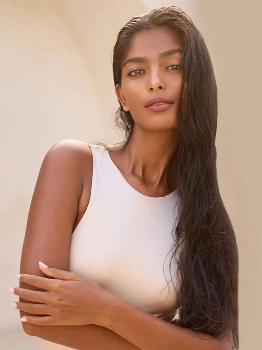 A portrait of a woman with long dark hair wearing a white sleeveless top, crossing her arms over her chest to subtly highlight the smoothness and tone of her upper arms. - Arm Lift in Arizona