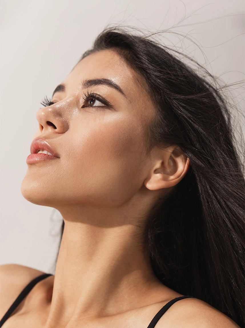 A profile view of a woman with dark hair looking up toward the light, highlighting her sculpted cheekbones and the defined, slender contours of her lower face and jawline. - Buccal Fat Removal in Arizona