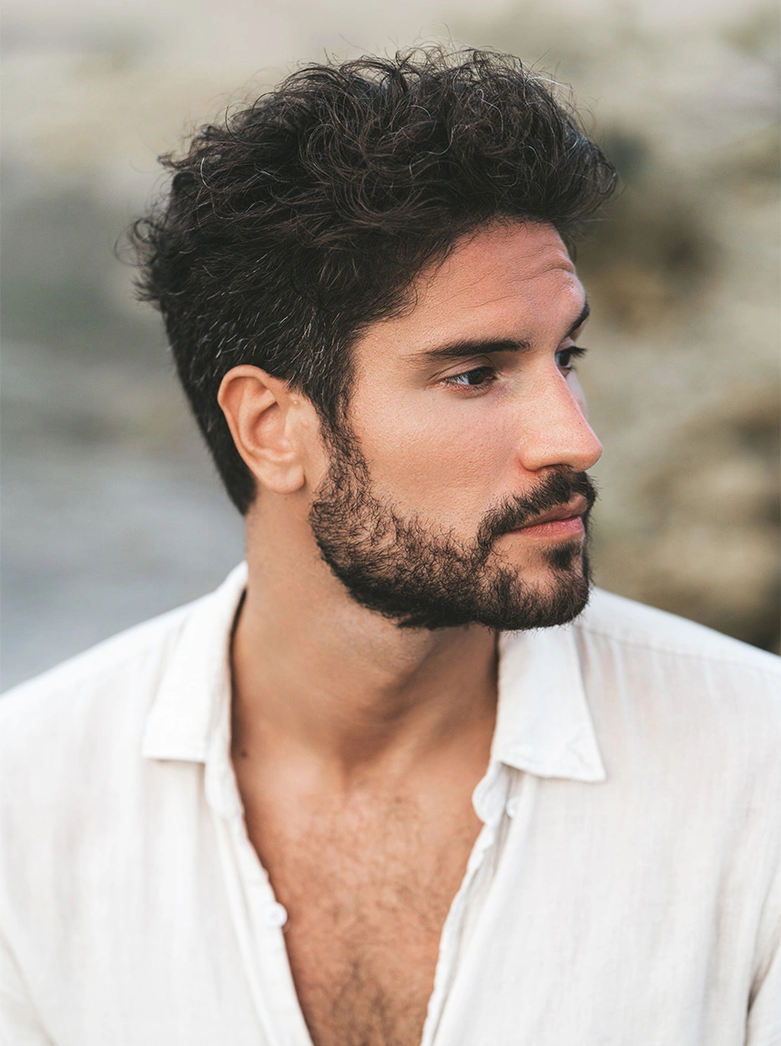 A portrait of a man with curly dark hair and a beard, wearing a white linen shirt. The image provides a clear view of his earlobe, showing a clean and natural appearance. - Earlobe Closure in Arizona