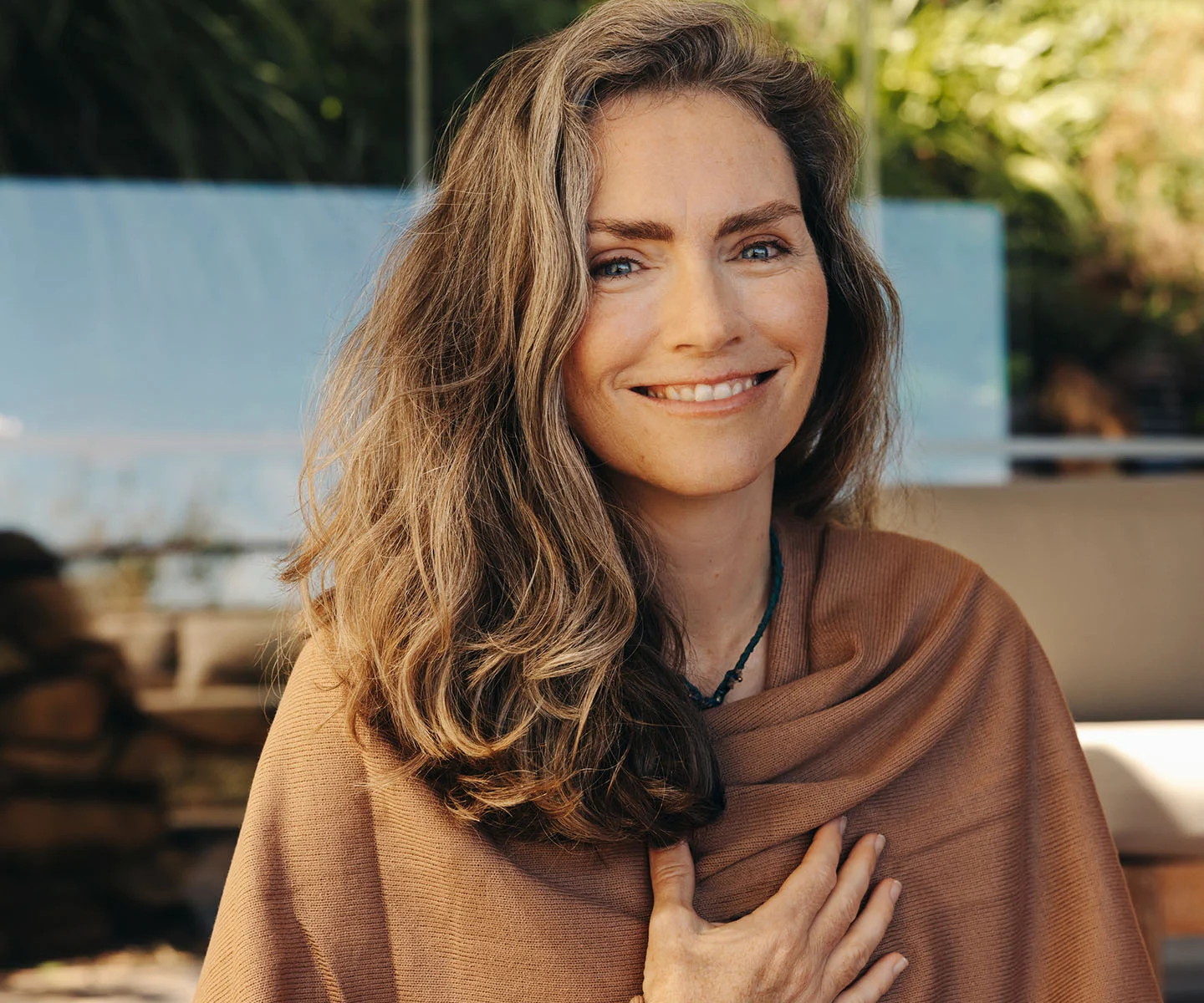 A lifestyle shot of a woman sitting comfortably and confidently on a modern, neutral-colored chair. she is wearing a flowing, light-colored dress and has a relaxed, happy expression while looking slightly off-camera. The environment is a bright, upscale lounge with soft textures and ample natural light, evoking a feeling of wellness and ease. - Emsella in Arizona