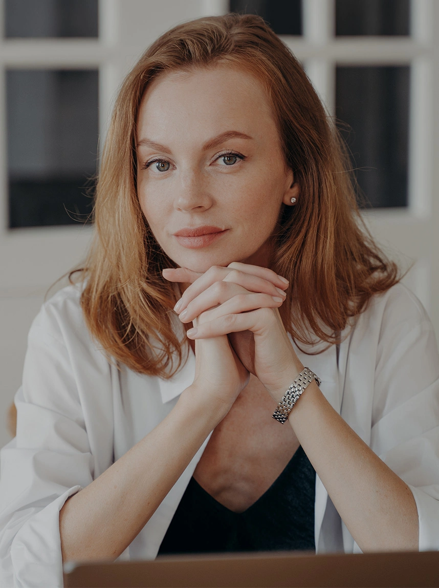 A woman with strawberry blonde hair wearing a white shirt sits with her hands clasped under her chin, featuring bright, open eyes and naturally elevated, symmetrical eyebrows. - Eyebrow Ptosis Repair in Arizona