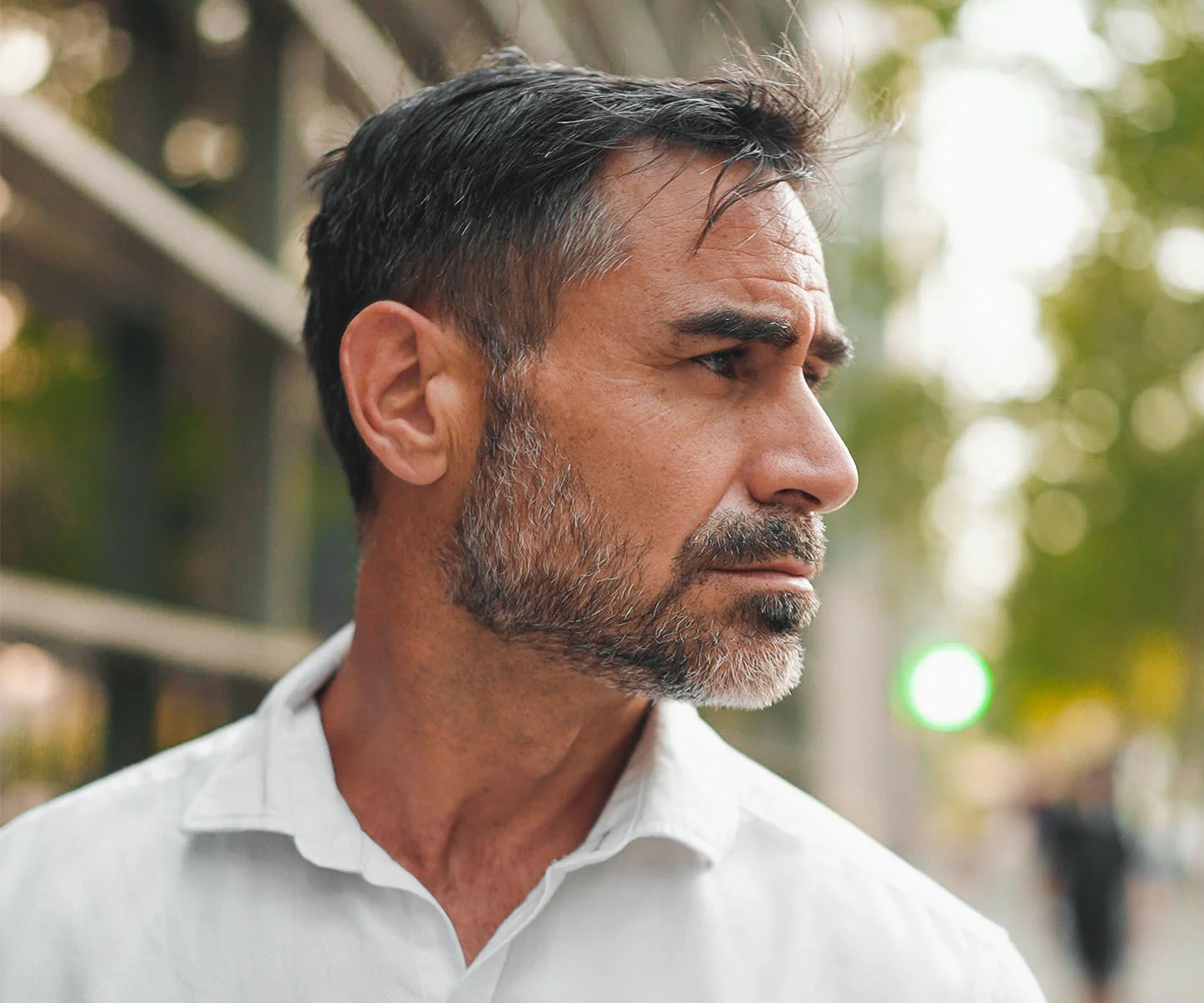 A medium close-up, profile view of a middle-aged man with short, salt-and-pepper hair and a neatly groomed beard. He is wearing a white collared shirt and is looking off-camera to the right with a thoughtful, serious expression. The background is a softly blurred outdoor setting featuring a modern building with metallic structures and lush green trees illuminated by natural sunlight. - Eyelid lesions in Arizona