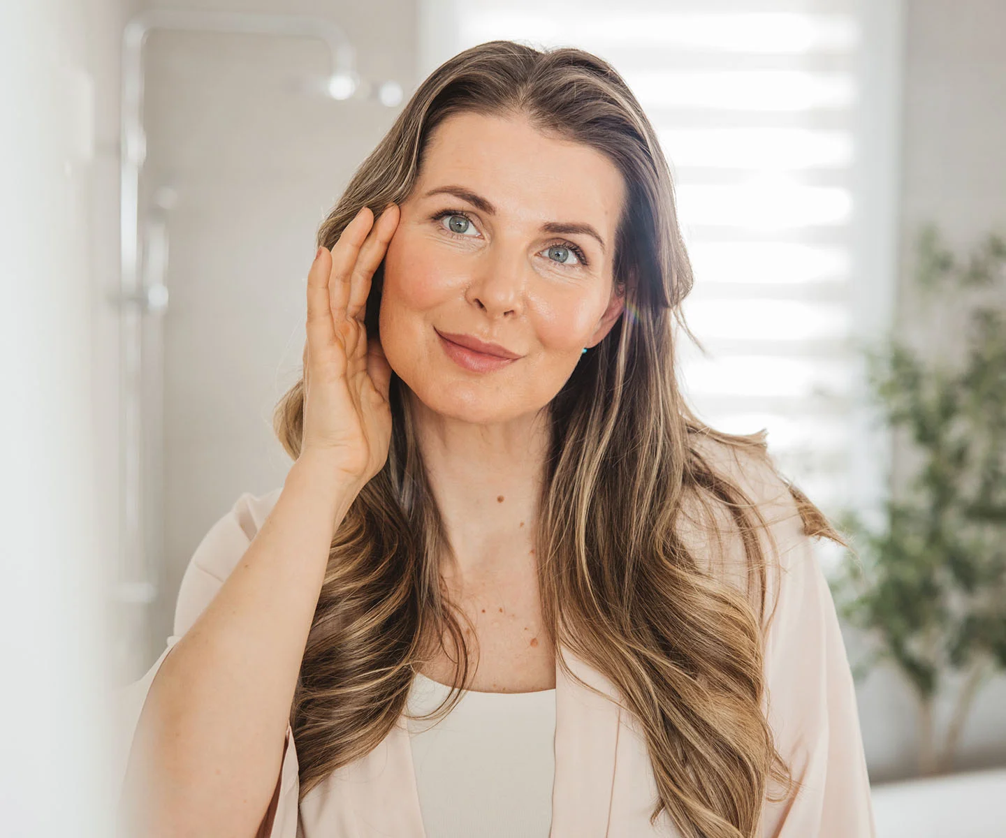 A portrait of a woman with long, wavy light-brown hair smiling warmly. She is gently touching her cheek, highlighting a symmetrical and natural eye area in a bright, soft-focus home setting. - Eyelid Misplacement in Arizona