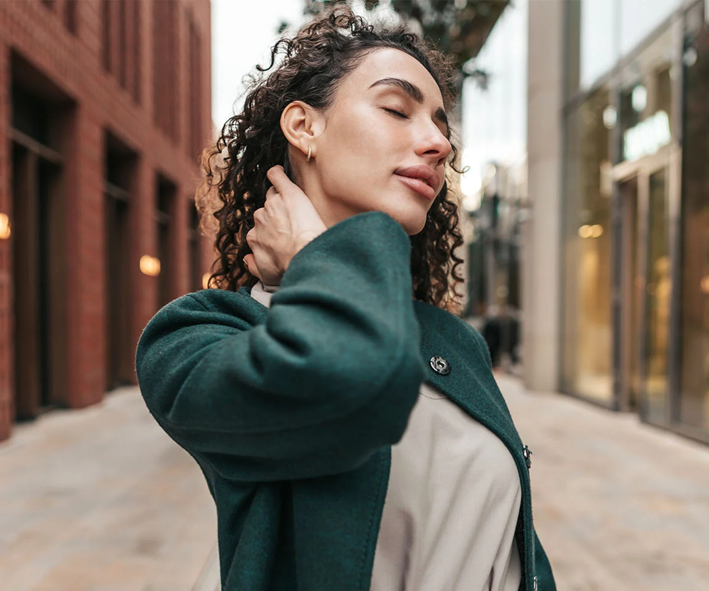 A woman with dark curly hair in a green coat tilts her head back with her eyes closed in an outdoor setting. The angle showcases full, youthful facial volume and smooth, balanced contours along her cheekbones and jawline. - Facial Balancing Fat Transfer in Arizona