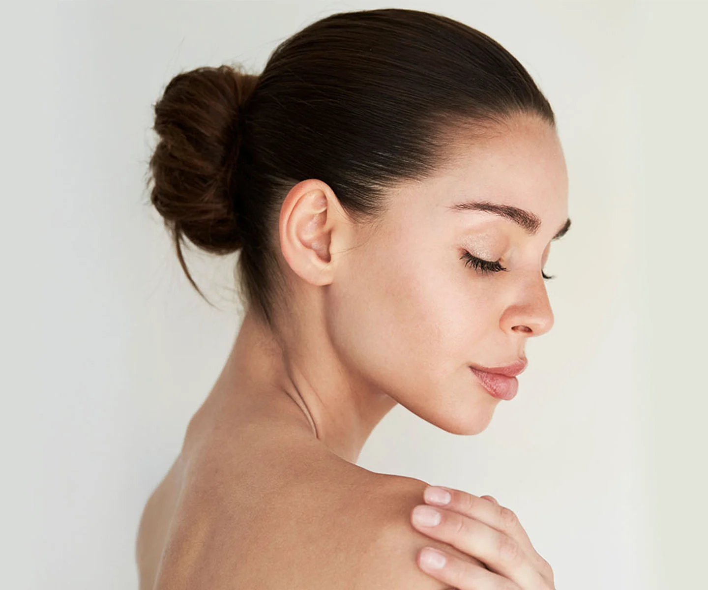 A woman with her hair in a sleek bun is seen from a side-profile view, touching her shoulder. The focus is on her smooth, clear skin and even complexion. - Facial Scar Revision in Arizona