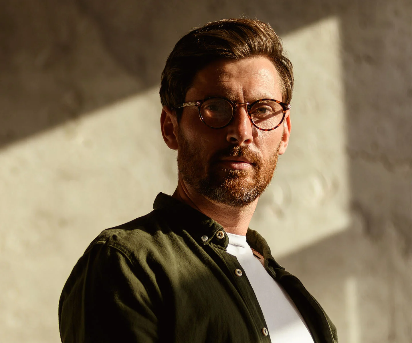 A portrait of a man with groomed brown hair and a beard, wearing tortoise-shell glasses and a dark green button-down shirt. Warm, angular sunlight illuminates his face, representing a sense of clarity, focus, and renewed vitality. - Hormone Replacement in Arizona