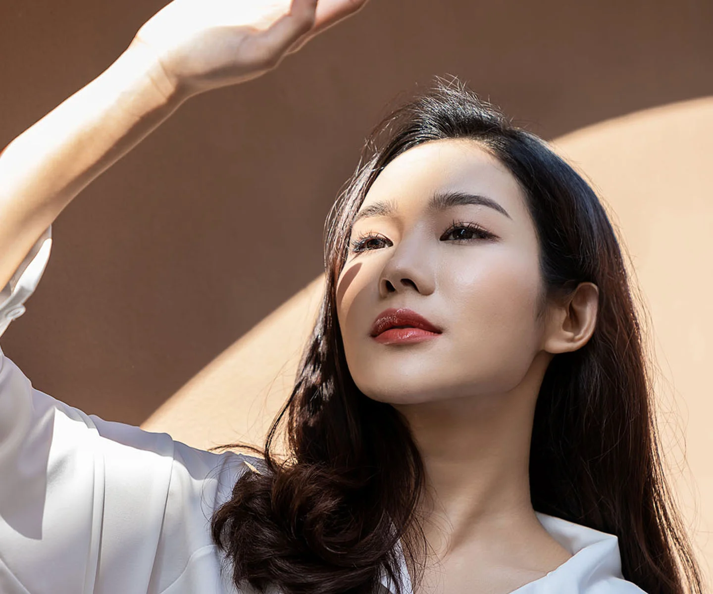 A woman with dark hair tilts her head back into the light, raising one hand to shade her eyes. The bright sunlight highlights a clear, radiant, and even skin tone. - Lasers in Arizona