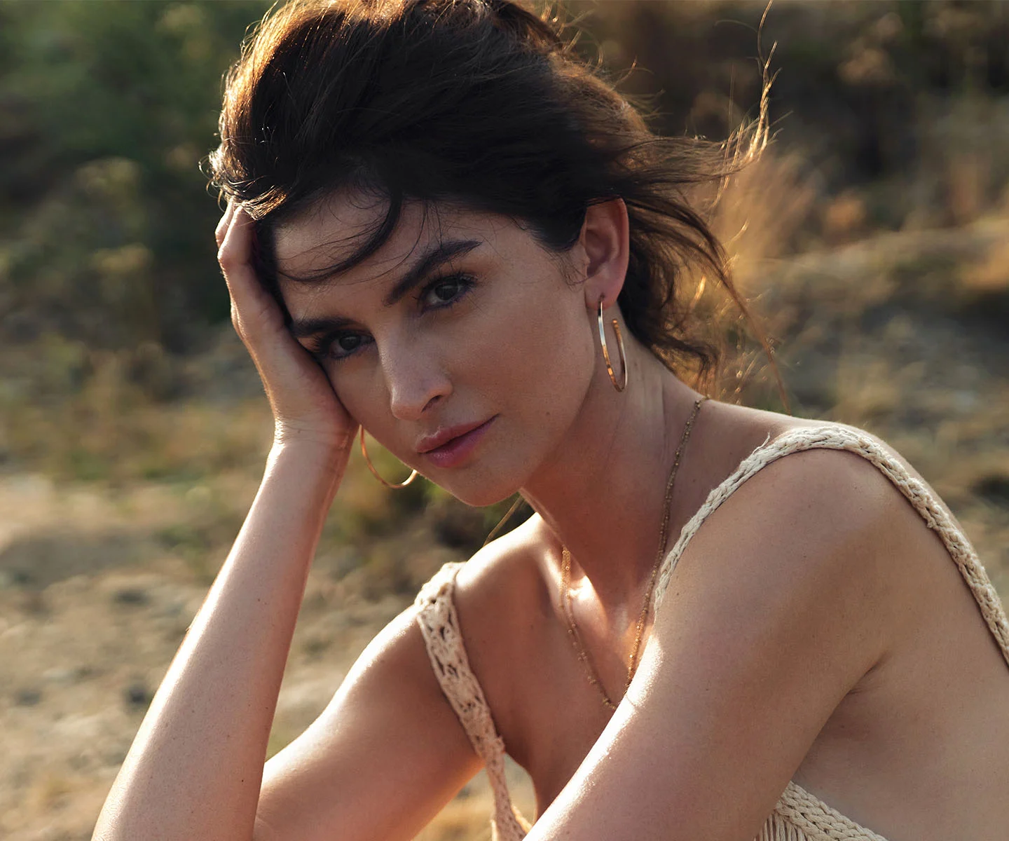 A woman with dark hair in a textured beige top looks directly at the camera while resting her head on her hand. The soft-focus outdoor setting emphasizes her bright, well-positioned eyes and healthy eyelid area. - Lower Eyelid Retraction in Arizona