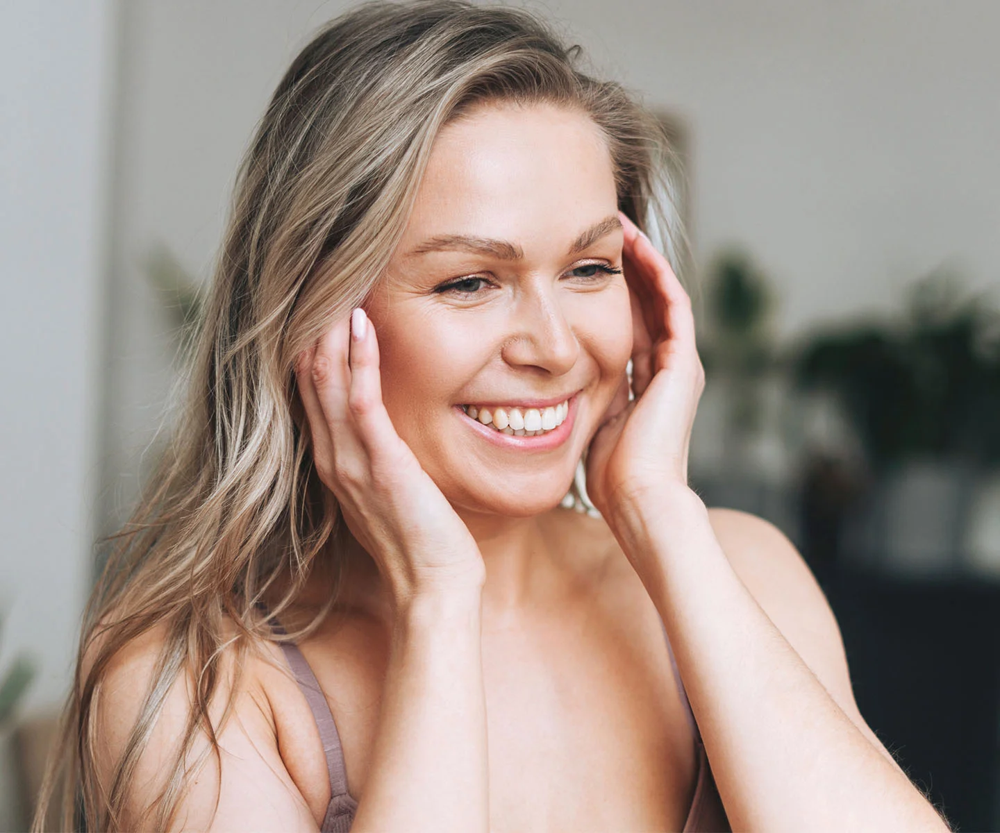 A woman with blonde hair smiles broadly while gently touching her temples, representing a sense of relief, comfort, and pain-free living. - Migranes in Arizona