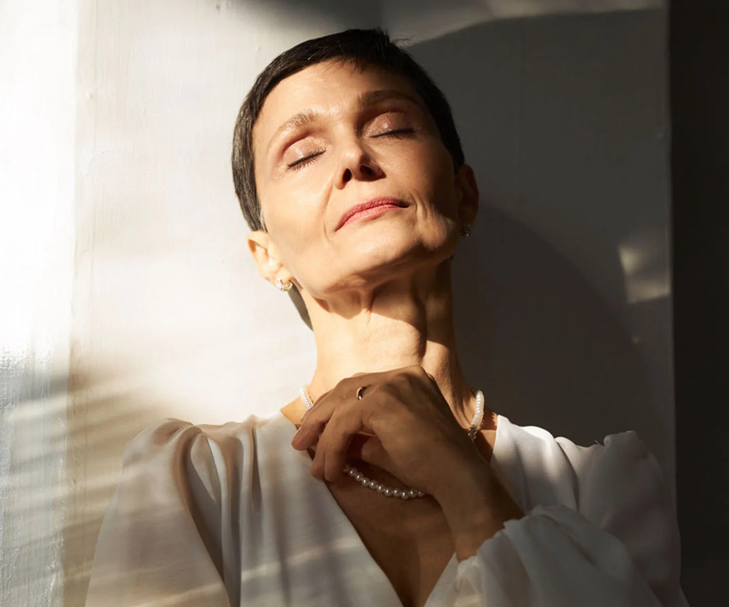 A woman with short dark hair and a pearl necklace closes her eyes in warm sunlight, tilting her head back to reveal smooth skin and a naturally restored facial appearance. - Mohs Reconstructive Surgery in Arizona