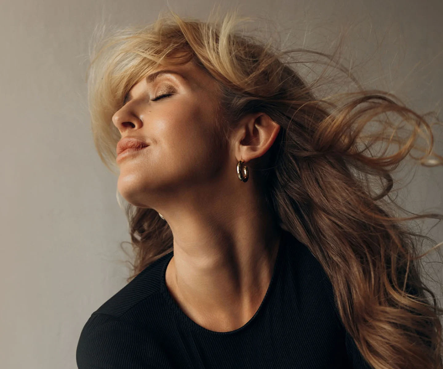 A profile view of a woman with her head tilted back and eyes closed, showcasing a sleek, tight neck profile and defined jawline. Her blonde hair flows behind her in a dramatic, wind-swept motion. - Neck Lift in Arizona