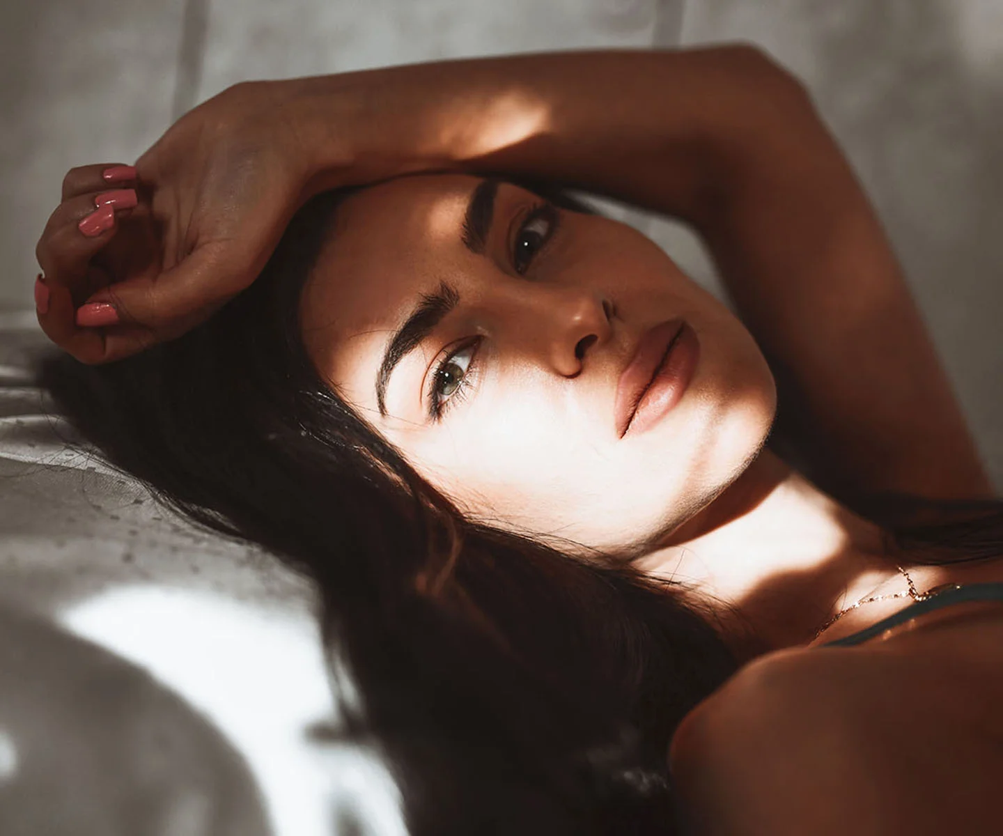 A woman with dark hair lies down in soft, dappled light, resting her head on her arm. The sunlight highlights the smooth, balanced bridge of her nose and refined facial symmetry. - Non-Surgical Nose Job in Arizona