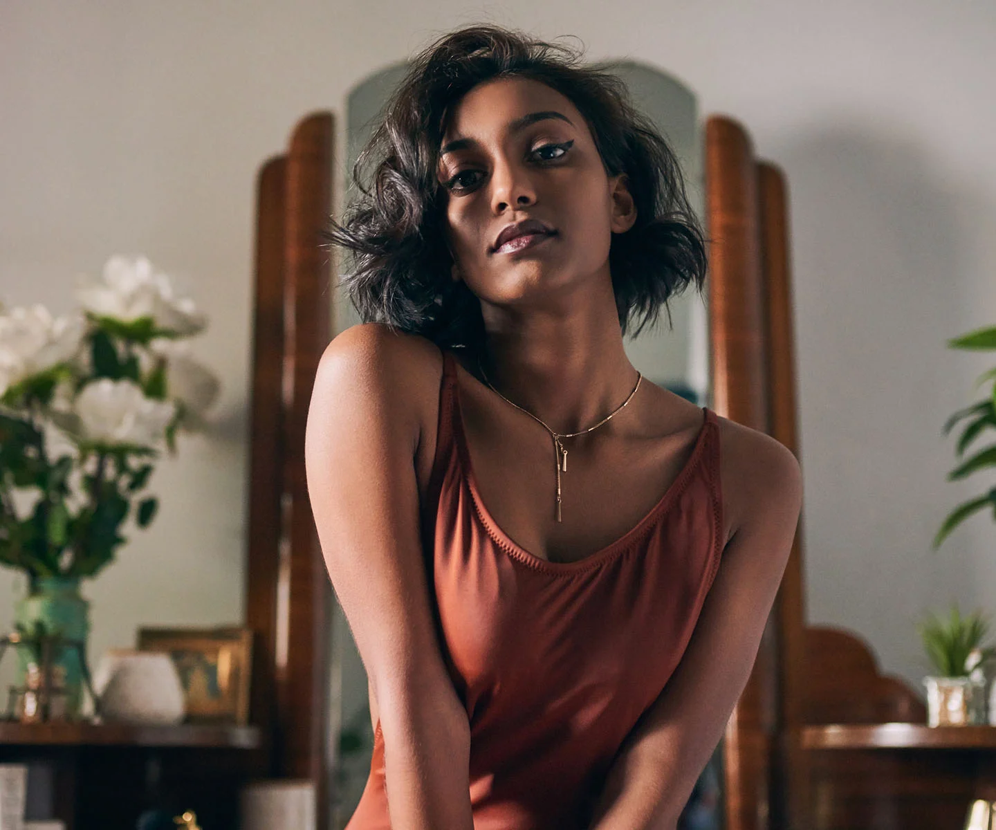 A portrait of a woman with short dark hair in a rust-colored bodysuit, posing in front of an elegant vintage mirror. The warm, sophisticated setting represents a comprehensive approach to beauty. - Overview in Arizona