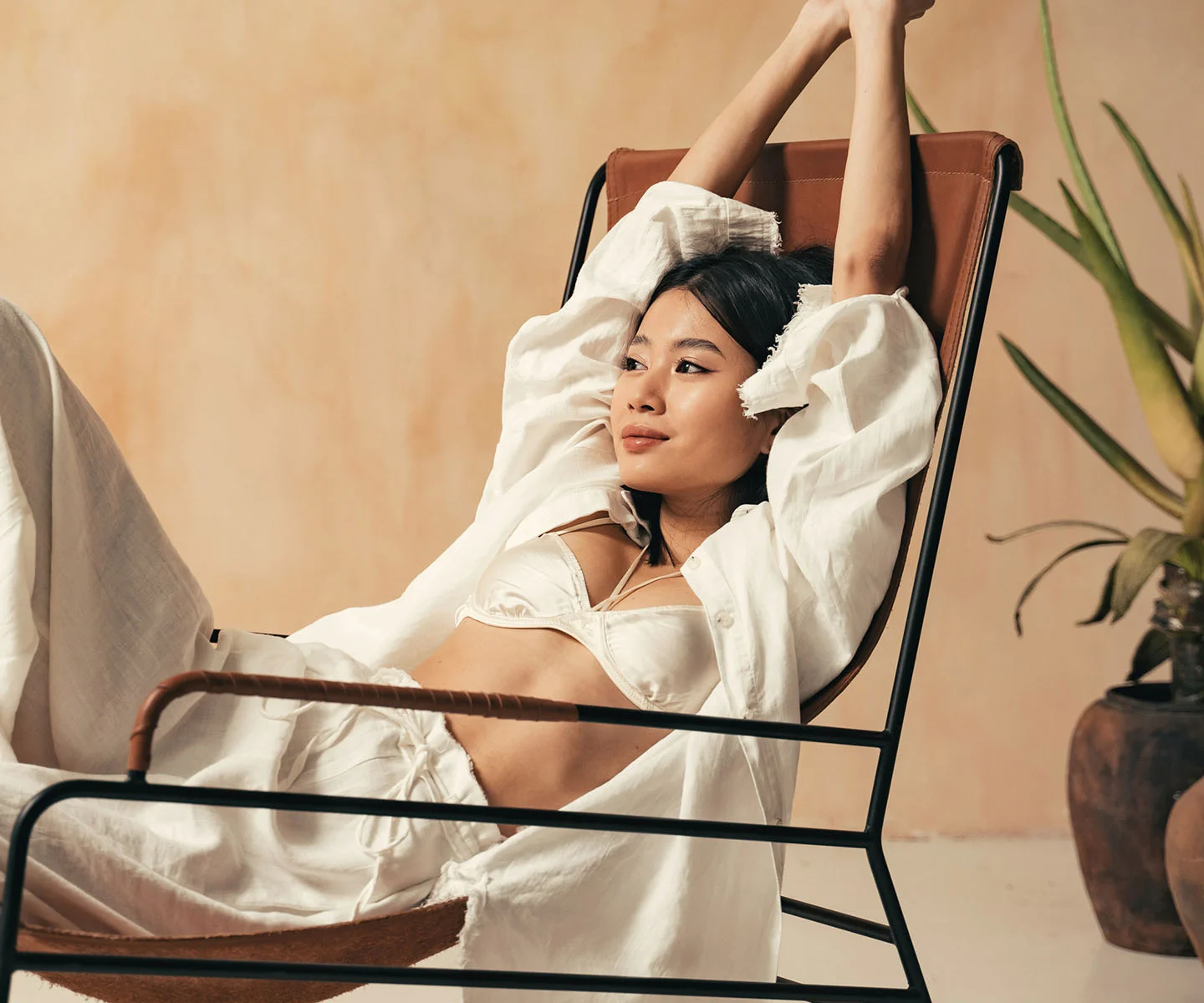 A woman in a white silk bra and trousers relaxes in a leather chair with her arms raised, showcasing a firm, youthful, and symmetrical upper body silhouette. - Renuvion for Breasts in Arizona