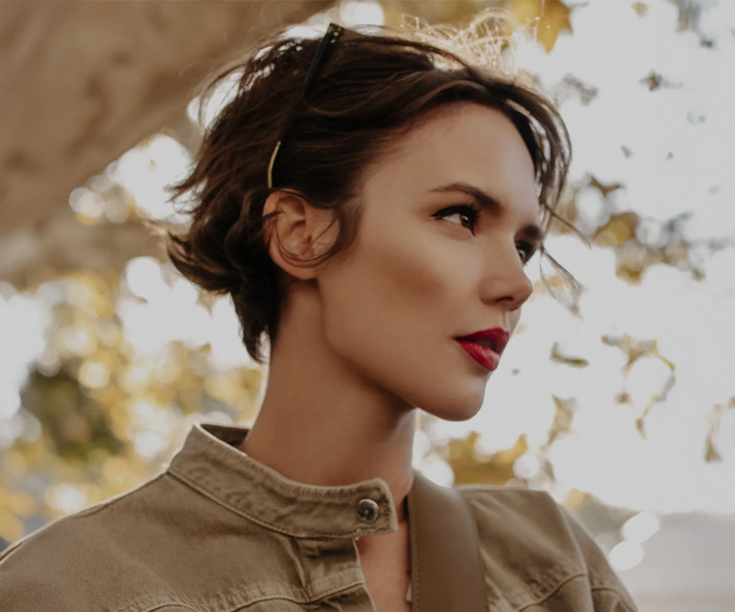 A side-profile portrait of a woman with short dark hair and bold red lipstick. The warm, sun-drenched background highlights her refined facial profile and smooth complexion.- Rhinoplasty in Arizona