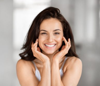 A joyful woman with a bright smile, holding her face with her hands, posed against a blurred background